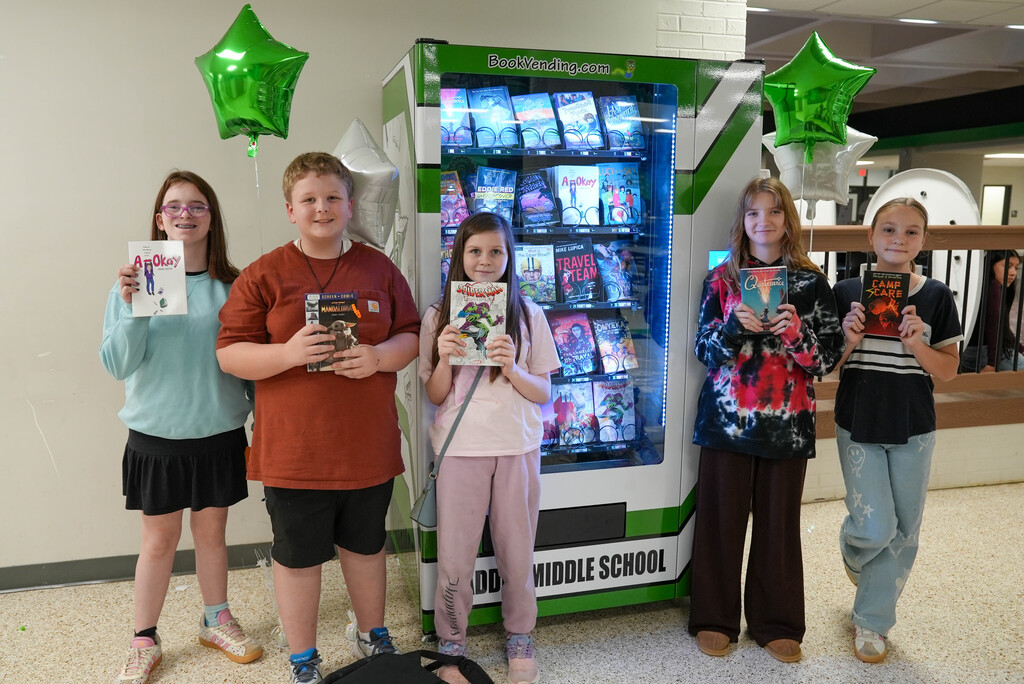 Five children stand in front of a green book vending machine, each holding a book. They wear casual clothing.