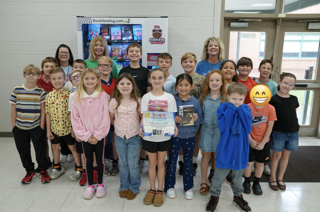 Group of children and two adults standing together for a photo with one holding a book.