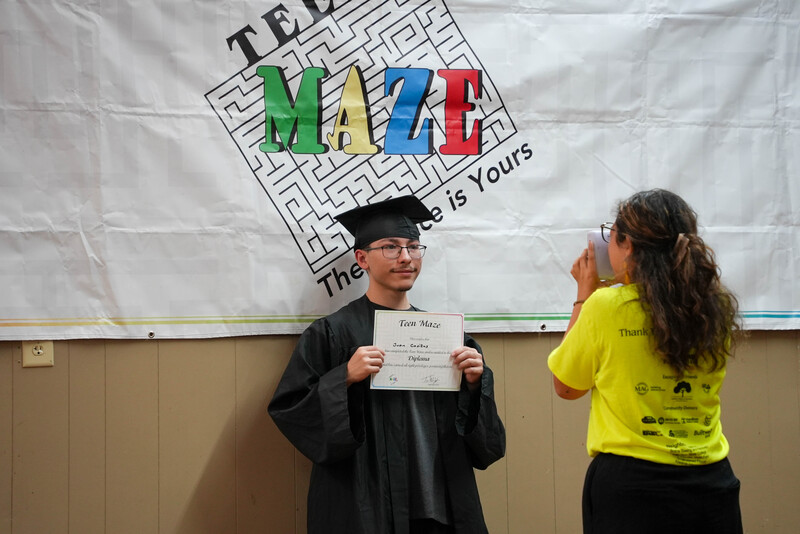Young man holds diploma in front of Teen Maze sign as volunteer takes his photo