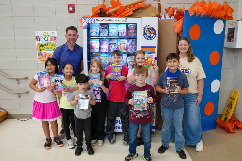 A group of students and teachers hold books up in front of book vending machine