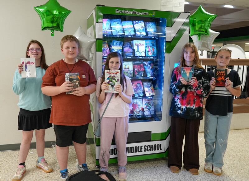 A group of students hold books up in front of book vending machine