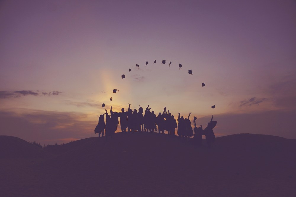 Graduating seniors stand outside on a sunny evening as the sunsets, tossing their mortar board caps in the air
