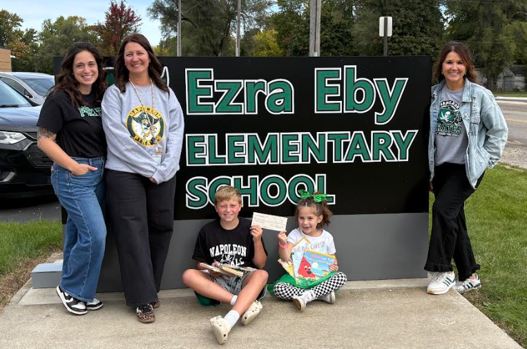 three women and two children holding books and check standing in front of Ezra Eby Elementary School sign