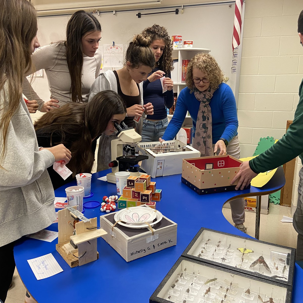 A high school science teacher stands with a group of middle school students as they look through a microscope.