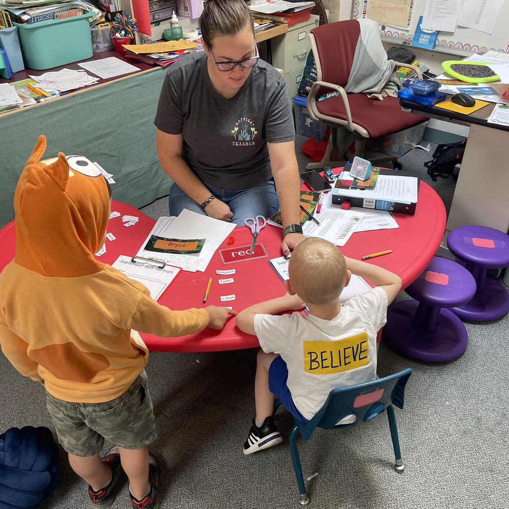 A pre-kindergarten teacher sits with two students at a table.