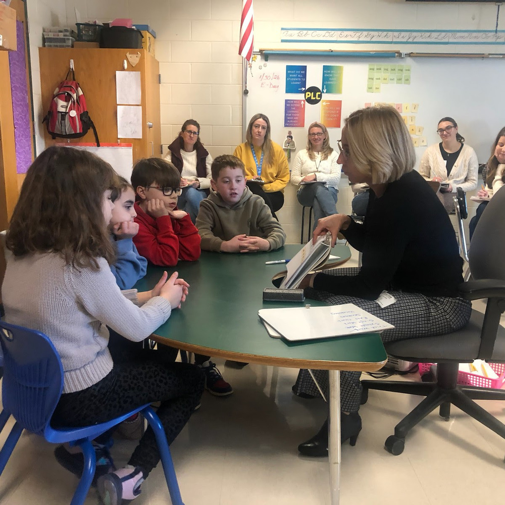 Elementary students sit at a table looking at a book with a teacher.