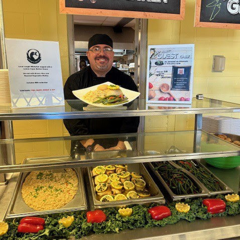 cafeteria staff member stands behind the serving line with a plate of food in front of him