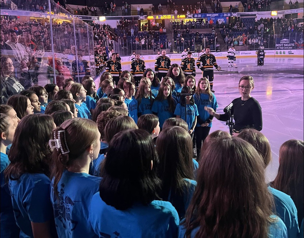 middle school students wear blue shirts and stand on the ice at the DCU center singing