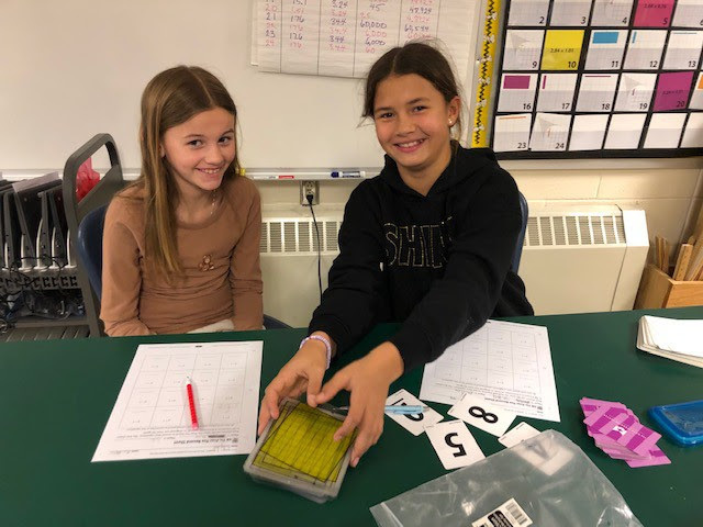 two students sit next to each other with paper and number cards in front of them