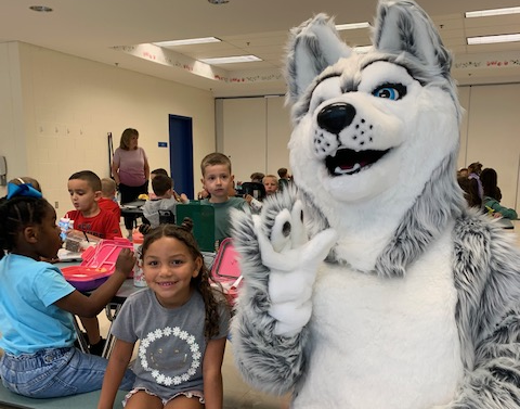 The MRE Huskey mascot sits with a smiling student and waves at the camera.