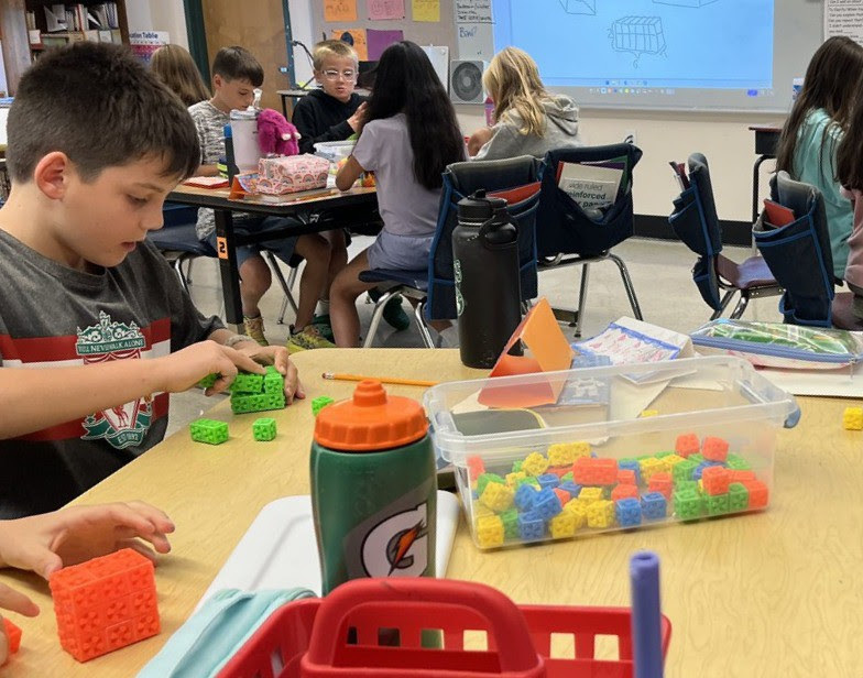 Elementary school students use colorful connecting blocks while sitting together at tables in small groups.