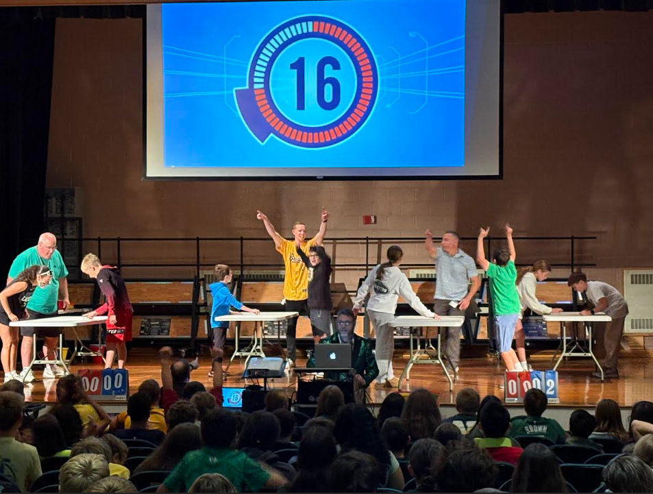 FSS Dean of Students Mark Schulze stands on stage with chearing students and teachers  while a timer counts down. Students sit in the auditorium seats watcing.