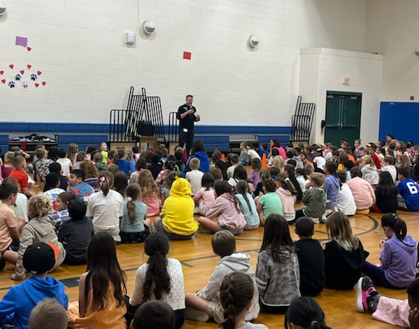 Students sit on the MRE gym floor while Principal Blanchard stands in front of the bleachers talking.