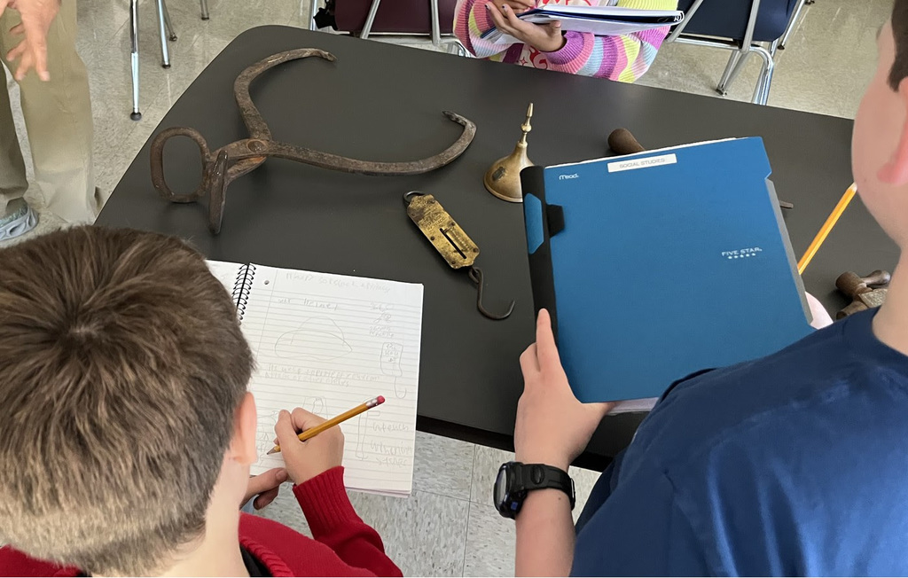 FSS sixth grader students write in notebooks as they examine artifacts on the table.