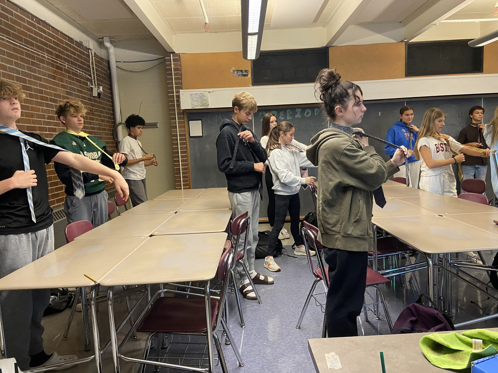 High school students stand in front of their desks putting on neckties.