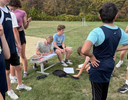 Students sit on a metal bench on a grass field doing schoolwork.