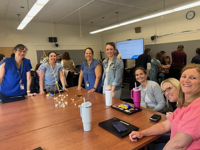 A group of teachers sits around a wooden table smiling. In the center is a toothpick and marshmallow tower.