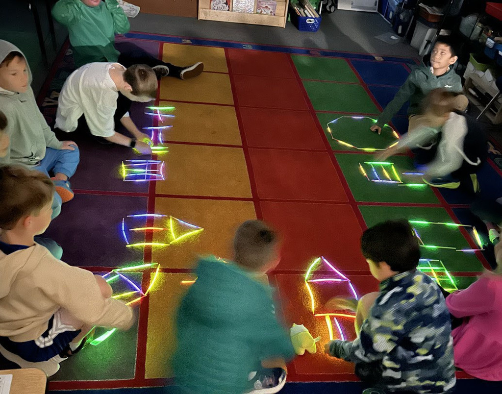 Elementary school students sit on a carpet with images that they've created out of glow sticks in front of them.