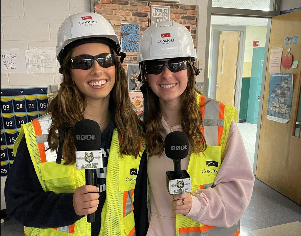 Two NRHS students wear Consigli construction hats and vests while holding Nashoba News microphones.
