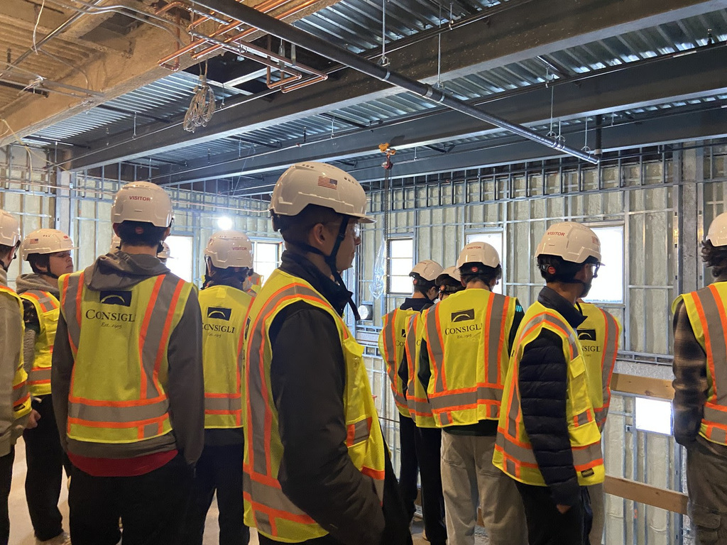 Students stand inside of a building being constructed wearing hard hats and high viz vests.