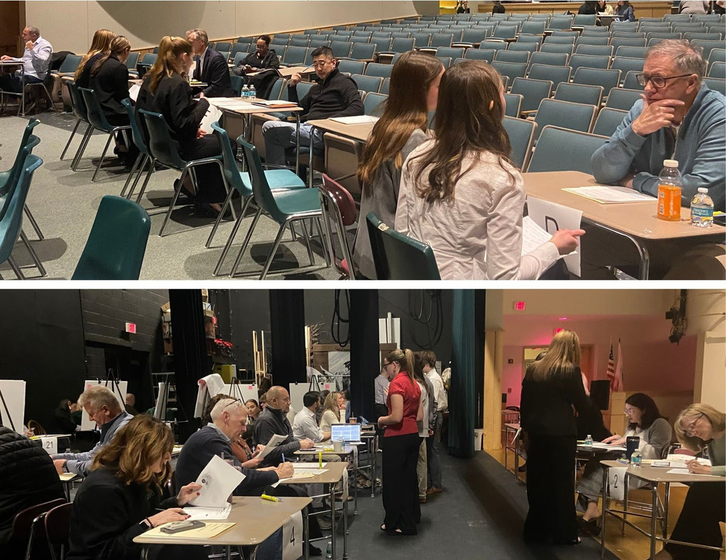 Judges sit at high school desks while high school students present projects to them.