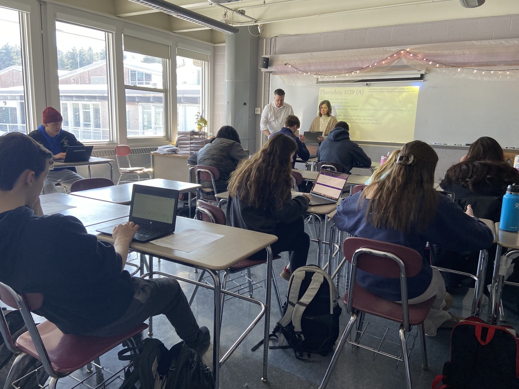 Students sit at their desks looking at chromebooks as two teachers stand at the front of the room.