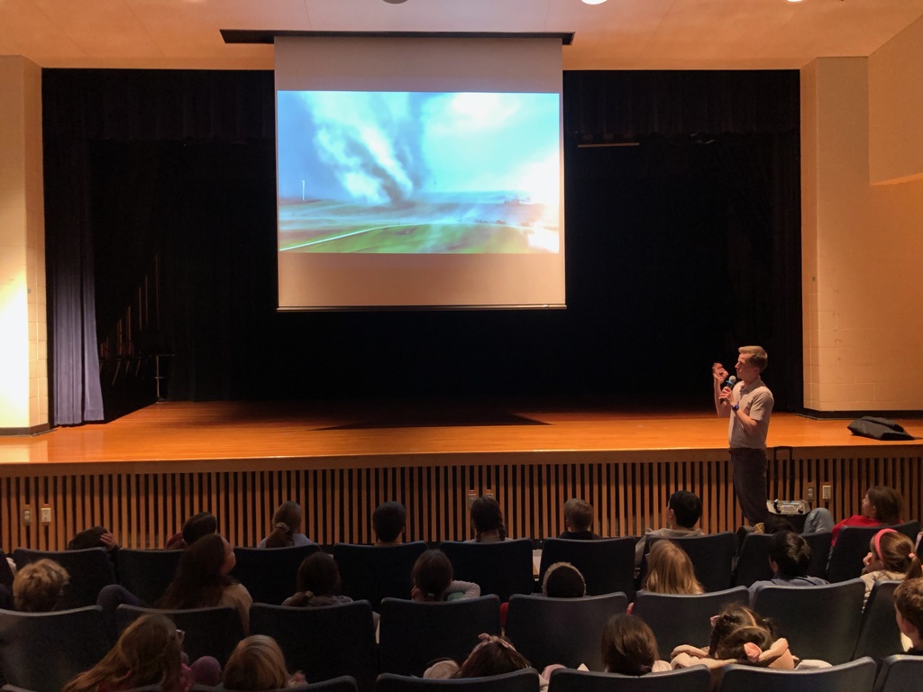 Students sit in the auditorium watching a meteorologist present while a video of a tornado is projected.