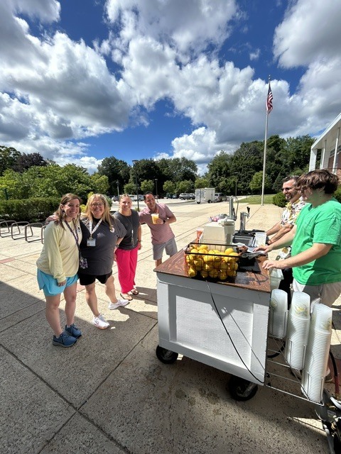 Teachers stand outside at a table with a basket of lemons and a juice press.