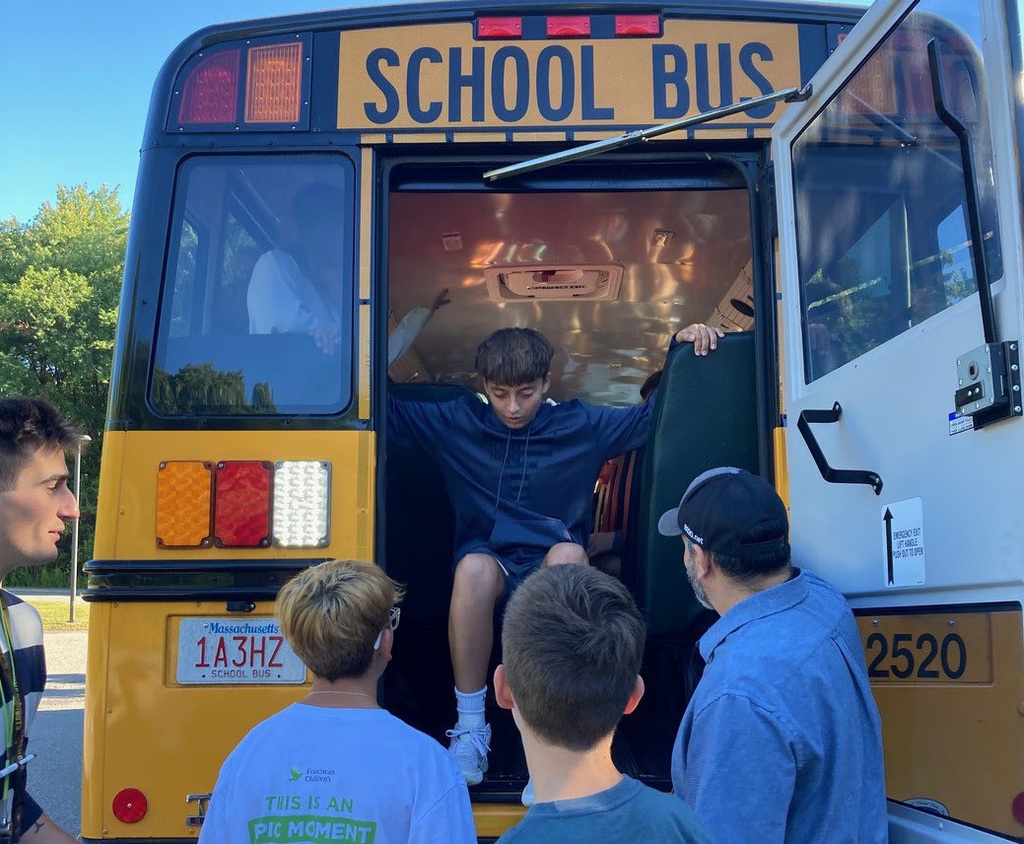 A student climbs out of the emergency exit of a bus during a bus drill while friends and teachers help.