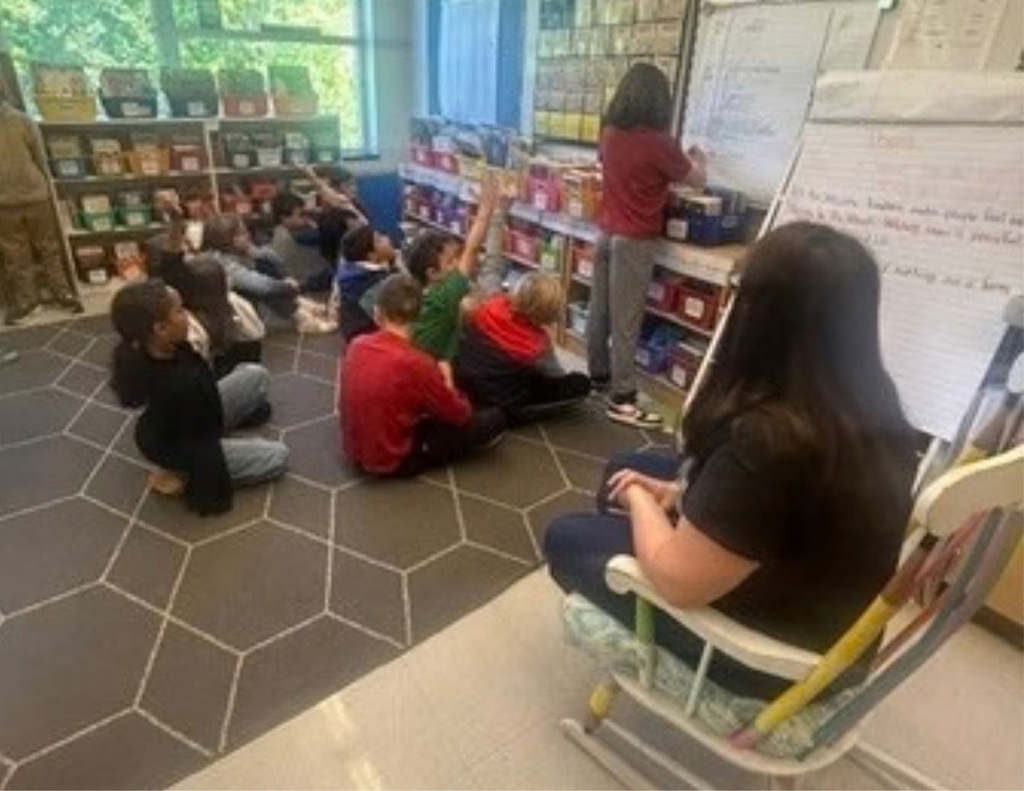 Elementary students sit on the rug watching another student complete a problem on the board.