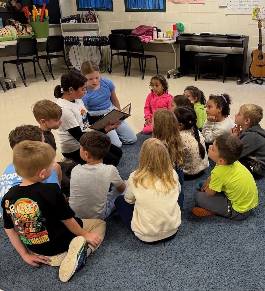 Elementary students sit on a rug in the music room as two older students lead a lesson alongside them.