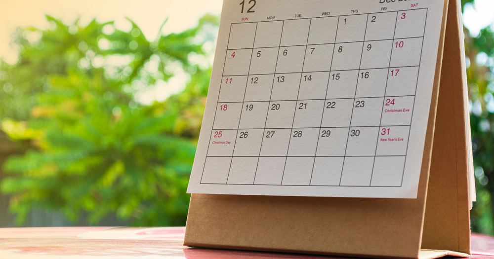 Paper desk calendar displaying the month of December, with holidays such as Christmas Day, Christmas Eve, and New Year’s Eve marked in red; calendar is set outdoors on a table with blurred greenery in the background.