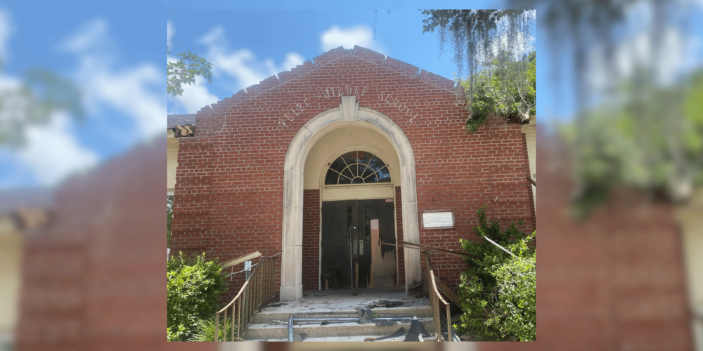 Front entrance of the historic Yulee School building during demolition preparations. The red brick facade remains standing, featuring a large stone archway surrounding the main entrance and the original "Nassau County High School" lettering above the doorway. Portions of the roof have been removed, exposing the building's deteriorated condition. Debris is visible on the front steps, and temporary railings line the entrance ramp. Green shrubs and oak trees with Spanish moss frame the building under a bright blue sky.