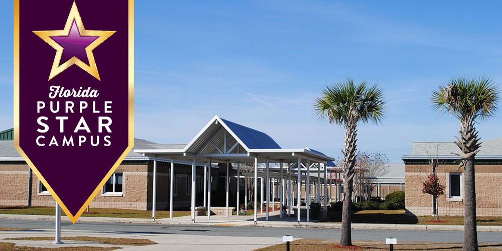 Exterior of Yulee High School with a covered walkway and palm trees in front of the building under a clear blue sky. A large purple and gold banner on the left displays the text “Florida Purple Star Campus.”