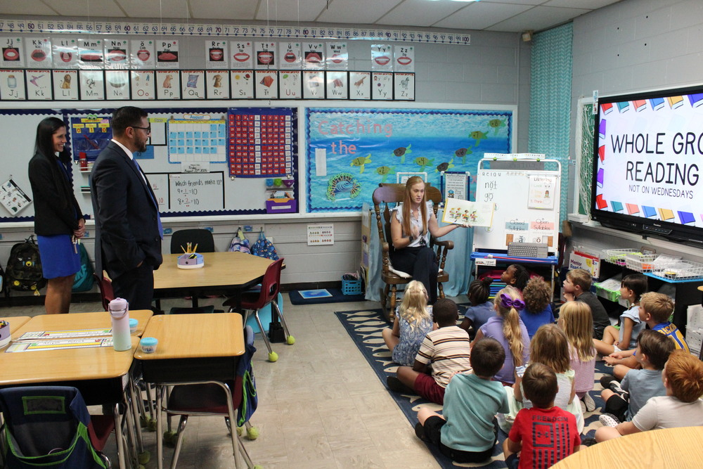 The Commissioner of Education and district leaders observe a teacher reading aloud to a group of elementary students seated on the floor during a whole group reading lesson.