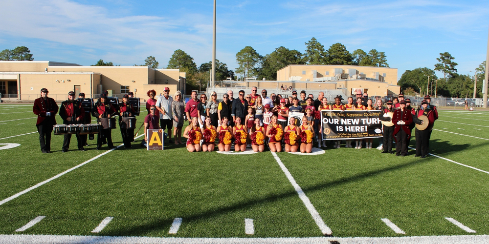 A completed group photo on the new turf field after the ribbon cutting. Students, cheerleaders, band members, and staff pose together, smiling behind a banner that reads, “Thank You, Nassau County. Our New Turf is Here! Funded by the One Mill.”