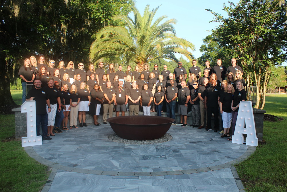 A large group of Nassau County School District leaders poses together outdoors during the annual Leadership Retreat. Everyone is wearing matching black shirts with the district logo, standing on tiered stone steps in front of a tropical palm tree. Large illuminated letters “1” and “A” are displayed on either side, symbolizing the district’s top-rated status.