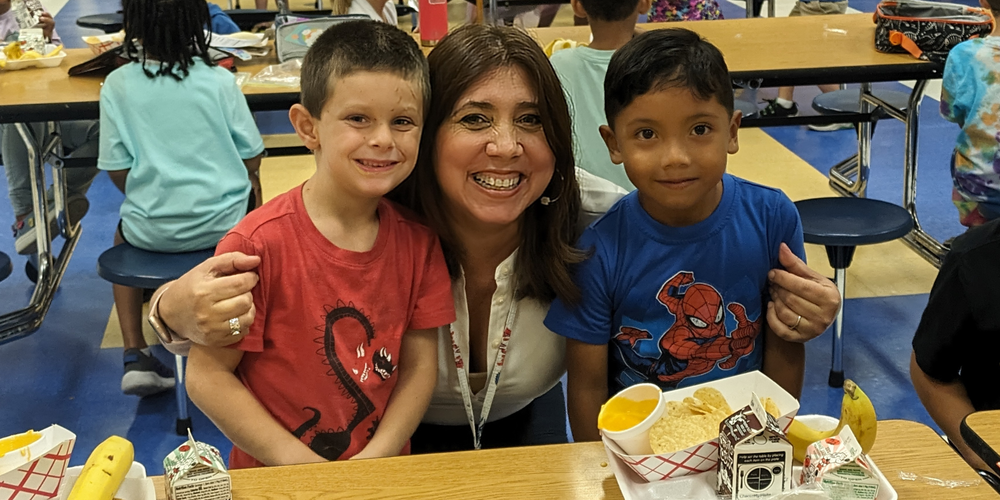 A smiling adult sits at a cafeteria table with two young elementary school boys during lunch. The child on the left is wearing a red dinosaur shirt, and the child on the right is wearing a blue Spider-Man shirt. Both are eating school lunch, which includes tortilla chips, cheese dip, a banana, and chocolate milk. The background shows other children seated at cafeteria tables.