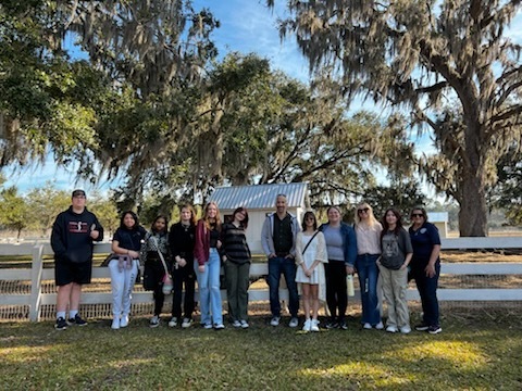 A group of students and adults pose for a group photo in front of a white wooden fence and a small structure under large moss-covered trees.