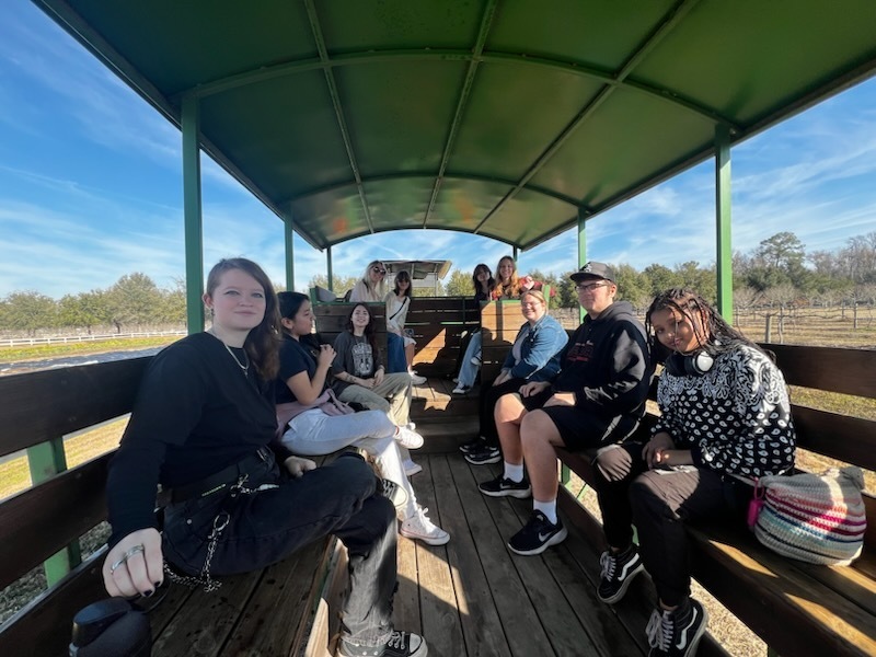 A group of students and adults sit on benches inside a green-roofed open-air wagon during a sunny ride around a rural property.