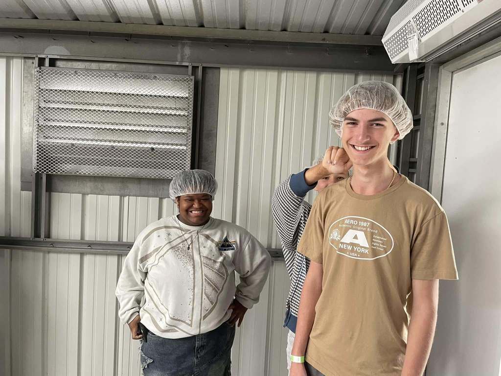 Two students smile while wearing hairnets inside a metal-paneled facility, with a third person playfully pointing at one of them from behind.