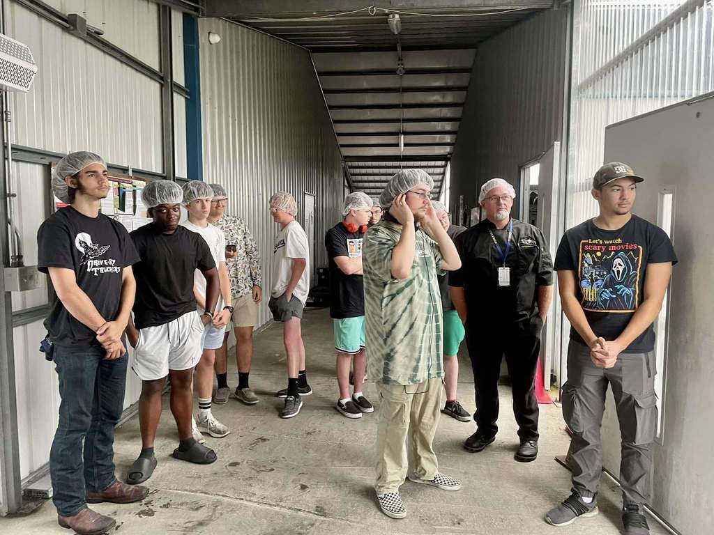 Students and adults wearing hairnets wait in a corridor outside a processing or facility room, some standing casually and others listening attentively.