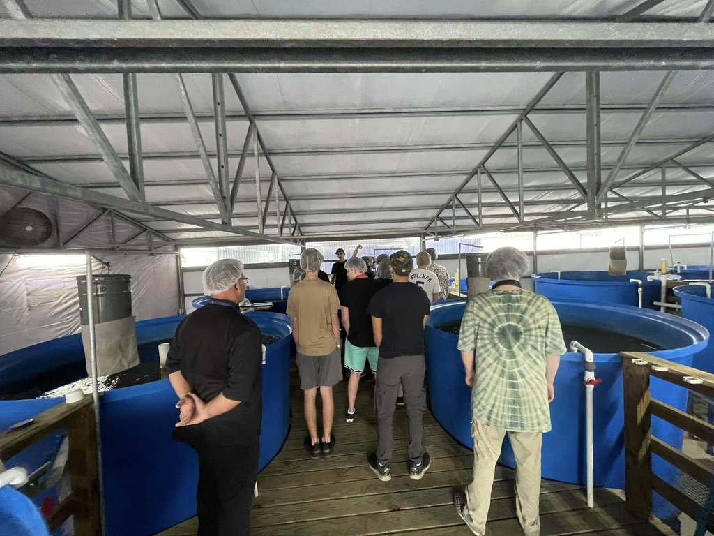A group of students and adults wearing hairnets stand inside an aquaculture facility surrounded by large blue circular fish tanks, listening to a guide.