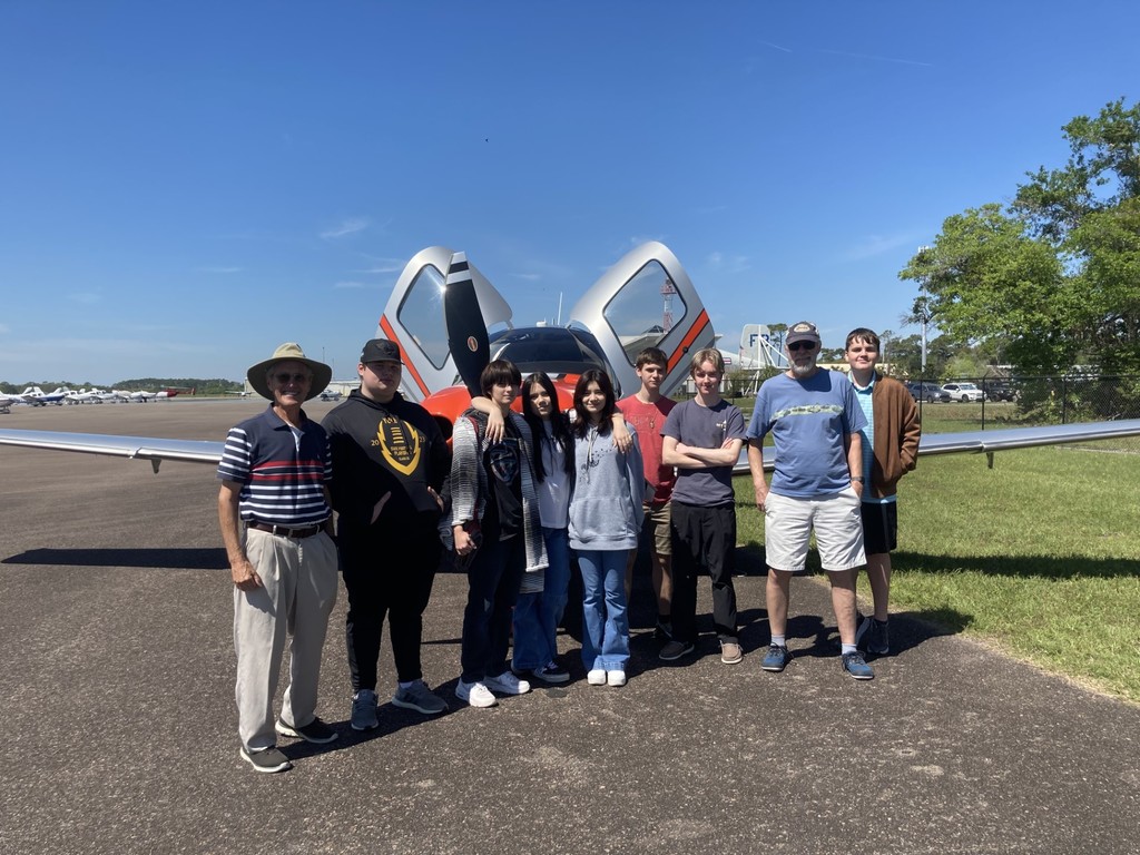A group of students and two adult instructors stand smiling in front of a white and red aircraft with both doors open on a sunny day at the airport.