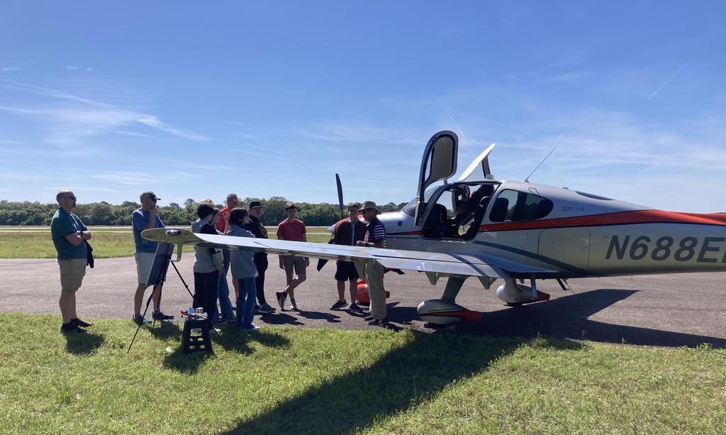 A different angle of an instructor providing aviation instruction near the aircraft’s nose, with a group of students and adults gathered around, watching attentively.
