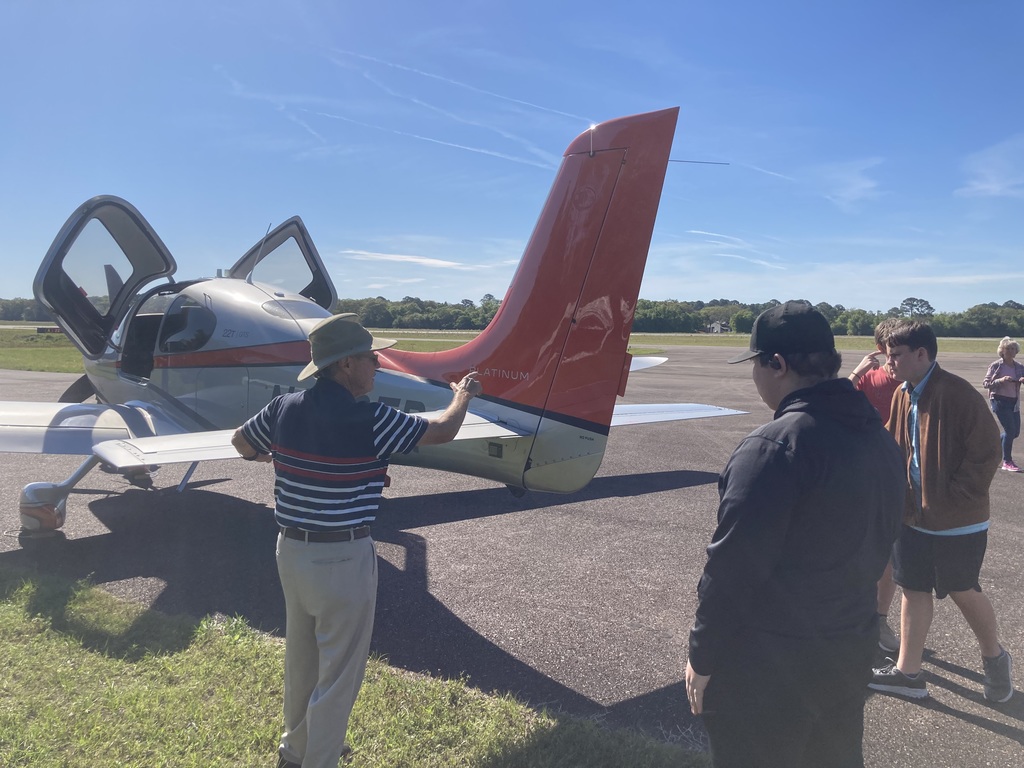 An aviation instructor wearing a wide-brim hat gestures toward the tail section of a small aircraft while a group of high school students listens attentively on the tarmac.