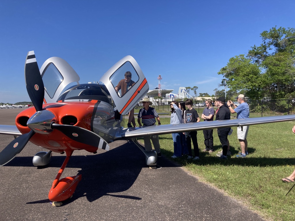 A high school student climbs into the side of a small aircraft as other students and adults stand nearby, observing and taking photos.