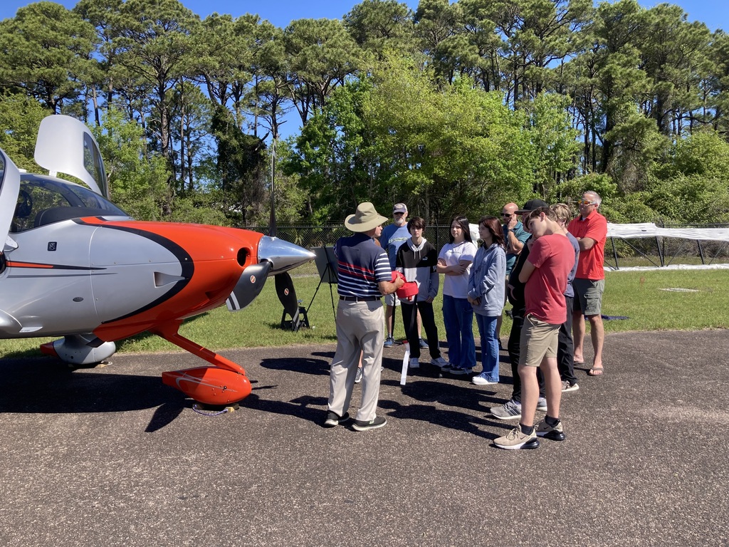An aviation instructor leads a lesson near the nose of a red and gray airplane while a group of students and adults listen and observe closely.