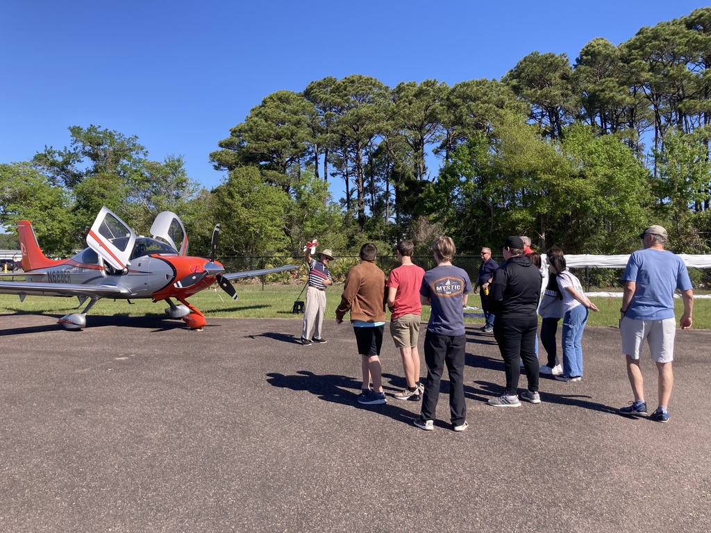 An instructor stands a short distance from the plane, holding up a red model airplane while explaining a concept to a group of students and adults.