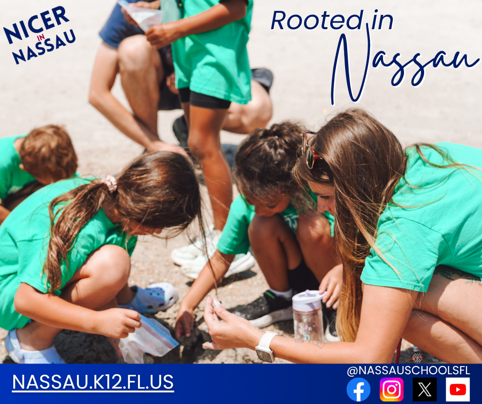 A teacher kneels in the dirt alongside a group of elementary students in matching green shirts, helping them examine something small during an outdoor learning activity.